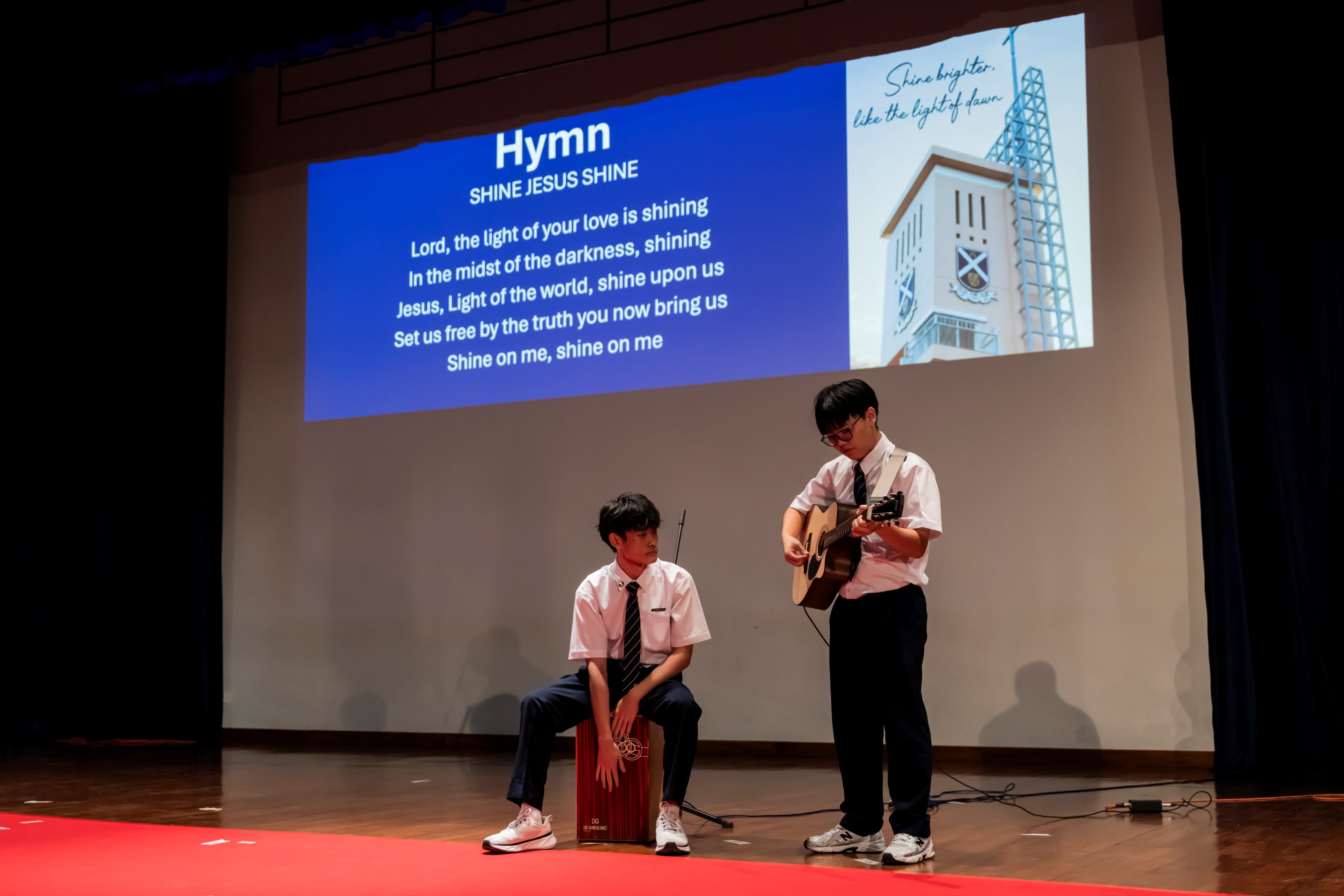 A stage performance comprising of 2 students in SAJC's school uniform playing a cajon and acoustic guitar respectively.